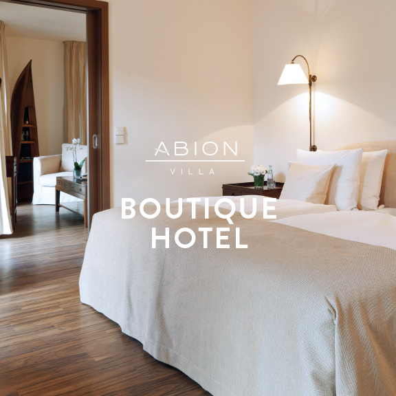 Foto: Hotelsuite der ABION Villa, Blick auf das Kingsize-Bett und den Durchgang in den Wohnbereich - Logo der ABION Villa und Aufschrift "BOUTIQUE HOTEL"