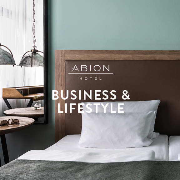 Foto: Hotelzimmer, Ausschnitt Bett und Holzmöbel - Logo ABION Hotel und Aufschrift "BUSINESS & LIFESTYLE"