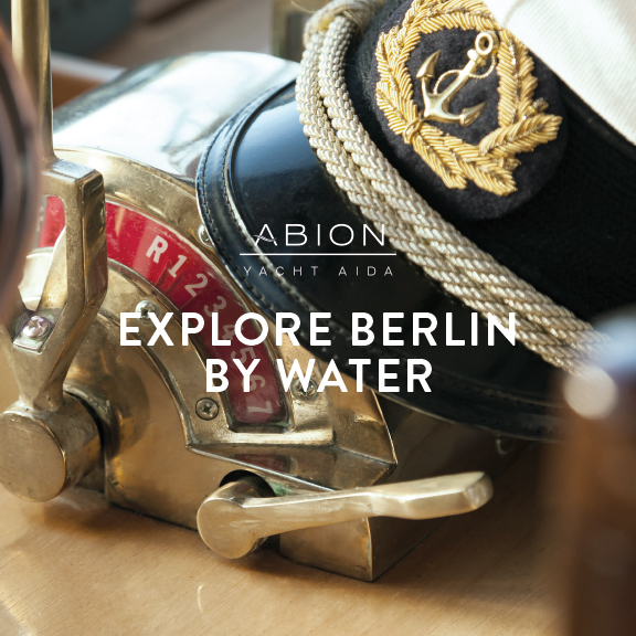 Foto : Yacht AIDA, goldener Steuerhebel und Capitainsmütze; Abion Yacht AIDA Logo und Aufschrift "EXPLORE BERLIN BY WATER"